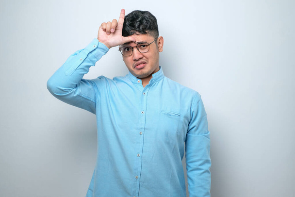 Young handsome man wearing casual shirt standing over white isolated background making fun of people with fingers on forehead doing loser gesture mocking and insulting. - Photo, Image