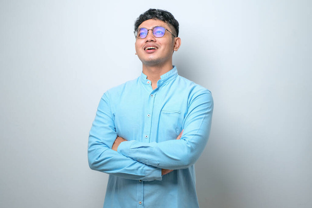 Portrét mladého pohledného asijského muže v ležérní košili a brýle stojící s rukama zkříženýma a usmívající se na kameru izolované přes bílé pozadí - Fotografie, Obrázek
