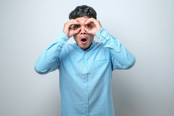 Young handsome man wearing casual shirt doing ok gesture like binoculars sticking tongue out, eyes looking through fingers. crazy expression over white background - Zdjęcie, obraz