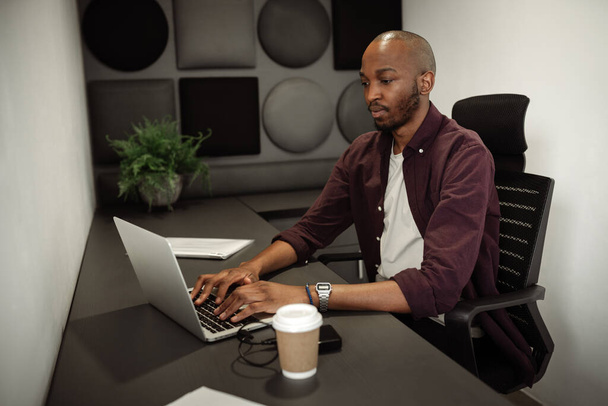 Afrykański biznesmen siedzi w biurze i pisze na laptopie. Niezależny przedsiębiorca pracujący przy stoisku biurowym - Zdjęcie, obraz