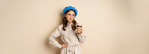 Όμορφο χαρούμενο κορίτσι πίνοντας καφέ takeaway από καφέ και χαμογελώντας, ποζάροντας με ένα φλιτζάνι ποτό, μπεζ φόντο. - Φωτογραφία, εικόνα