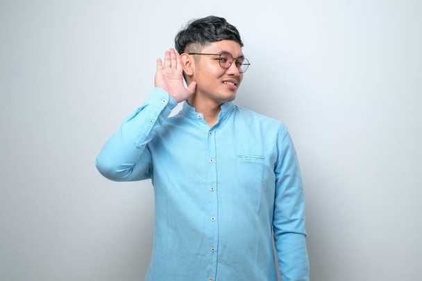 Mladý pohledný Asiat v neformální košili, stojící nad osamělým bílým pozadím a usmívající se s rukou přes ucho, naslouchající drbům a drbům. Koncept hluchoty. - Fotografie, Obrázek