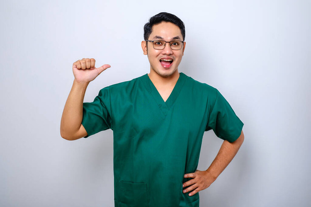 Professionelle selbstbewusste männliche asiatische Arzt, Krankenschwester in Peelings zeigt auf sich selbst und lächelt, Garantie, dass er weiß, was tun, weißer Hintergrund - Foto, Bild