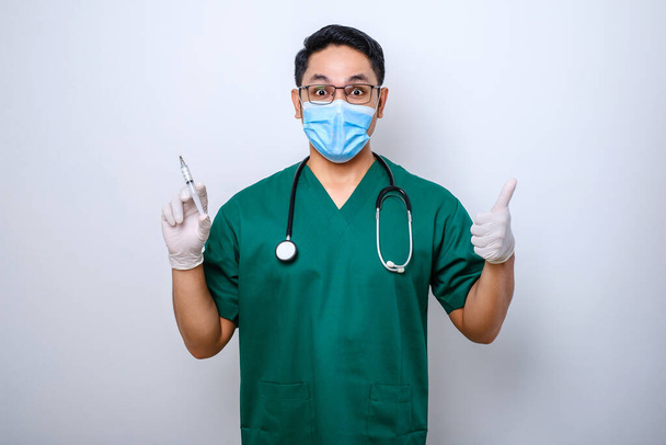 Aangename lachende Aziatische mannelijke arts, arts met medisch masker en handschoenen, duim omhoog, spuit vasthouden met vaccin geïsoleerd over witte achtergrond - Foto, afbeelding