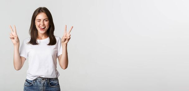 Cheerful cute brunette girl showing peace gestures and winking happy. - Foto, afbeelding