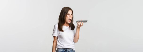 Arrabbiato ragazza bruna gridando pazzo durante la registrazione del messaggio vocale, essendo indignato, sfondo bianco. - Foto, immagini