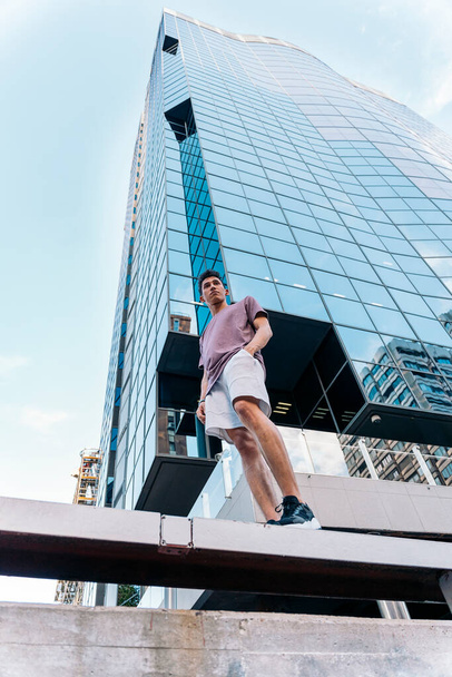 Cool young man wearing casual clothes posing and looking at front against modern building. - Zdjęcie, obraz