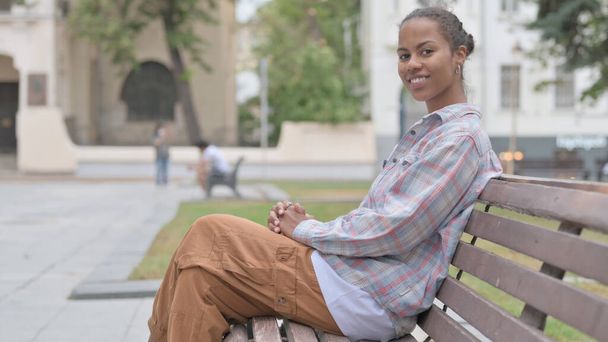 ベンチに座りながらカメラで微笑む若いアフリカ人女性 - 写真・画像