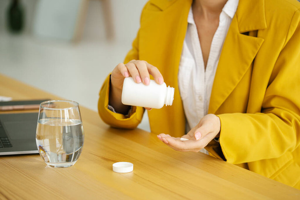 Close up of woman holding pill and glass of water in office in yellow suit - Фото, изображение