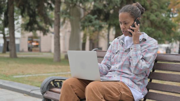 Arrabbiato giovane donna africana parlando al telefono e utilizzando il computer portatile mentre seduto all'aperto sul banco - Foto, immagini