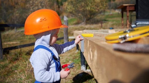Παιδί παιδί παίζει ως οικοδόμος στο εργοτάξιο. Παιδί ξυλουργός σε πορτοκαλί κράνος και μπλε φόρμα μάθησης για να χτίσει ξύλινο σπίτι πλαίσιο εξωτερική την ηλιόλουστη μέρα. Έννοια ξυλουργικής και εργαστηρίου. - Φωτογραφία, εικόνα