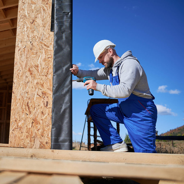 Male builder installing black corrugated iron sheet used as facade of future cottage, blue sky on background. Man worker building wooden frame house. Carpentry and construction concept. - Fotoğraf, Görsel