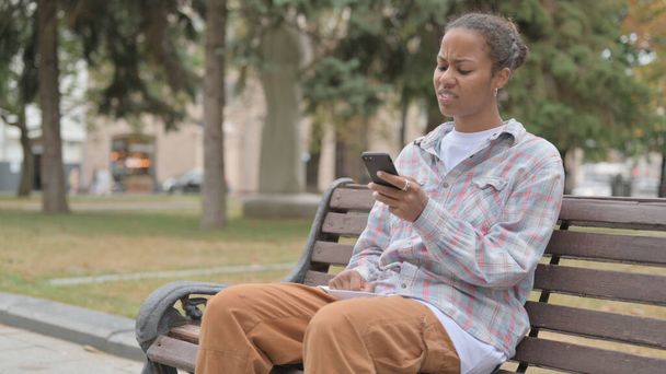 Молода африканка реагує на втрати на смартфоні сидячи на відкритому повітрі на лавці
 - Фото, зображення