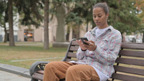 Молода африканка використовує смартфон сидячи на відкритому повітрі на лавці
 - Фото, зображення