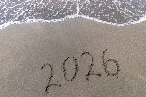 Κείμενο Καλή Χρονιά 2026 σε μια αμμώδη παραλία με ένα μικρό κύμα. Αφηρημένη φωτογραφία φόντου του επερχόμενου νέου έτους 2023 - Φωτογραφία, εικόνα
