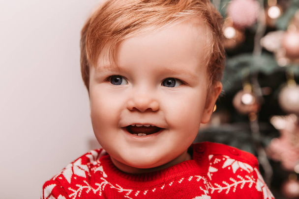 Młody dzieciak ubrany w czerwony ciepły sweter, siedzący obok choinki z zabawkami, uśmiechnięty. Świętowanie Nowego Roku lub Bożego Narodzenia w domu.  - Zdjęcie, obraz