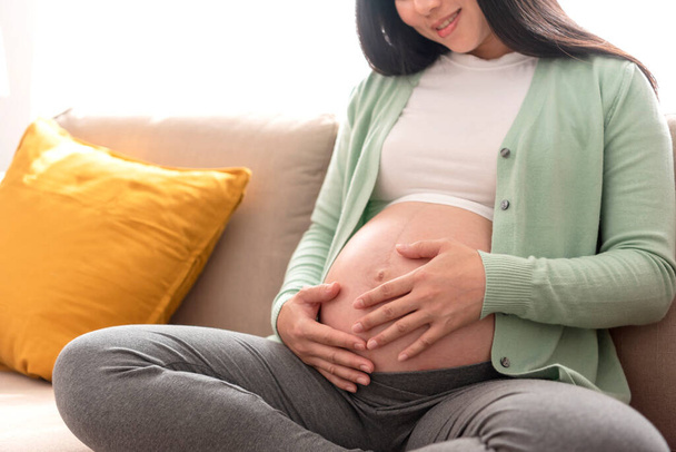 Молодая азиатская красивая беременная женщина ласкает руки на животе, сидя дома на диване. Беременность, материнство, подготовка и ожидание. - Фото, изображение