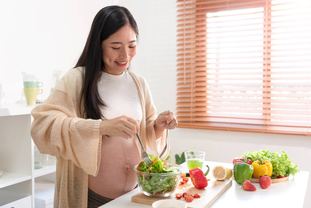 Glückliche asiatische schwangere Frau, die zu Hause Salat kocht, frischen grünen Salat macht, während der Schwangerschaft viele verschiedene Gemüsesorten isst, gesundes Schwangerschaftskonzept. - Foto, Bild