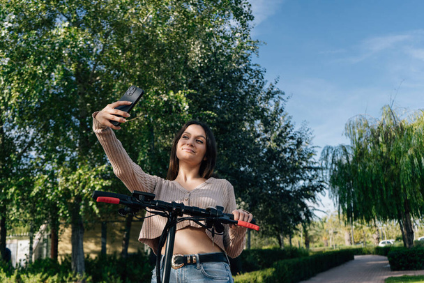 Elektrický skútr a mobilní zařízení. Mladá dívka používá chytrý telefon. Beru si selfie s elektrickým skútrem v městském parku. fotografie krajiny. - Fotografie, Obrázek