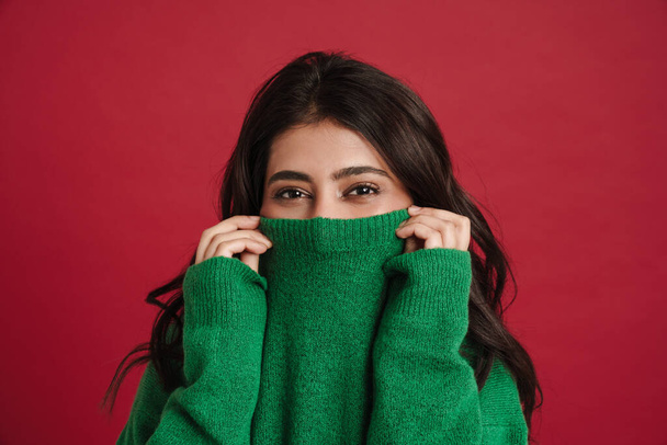 East asian woman hiding in sweater and looking at camera isolated over red background - Photo, Image