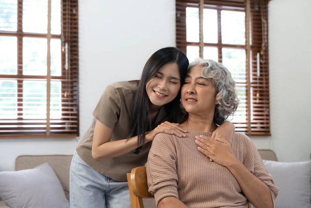 Loving adult daughter hugging older mother, standing behind couch at home, family enjoying tender moment together, young woman and mature mum or grandmother looking at each other, two generations.. - Photo, Image