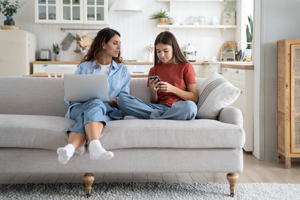 Friendly optimistic mother and daughter sits on sofa use modern electronic gadgets. Good-natured caring woman prompting teenage girl online dating precautions recommended reliable messenger apps - Fotoğraf, Görsel