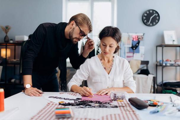 Competent male couturier with female assistant drawing sketches while designing exclusive accessories. Caucasian man and woman working together in modern atelier. - Photo, Image