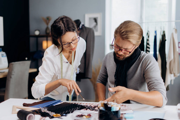 Dos diseñadores de moda caucásicos sentados juntos en un taller moderno y examinando los detalles para la futura colección. Proceso creativo en el taller. - Foto, imagen