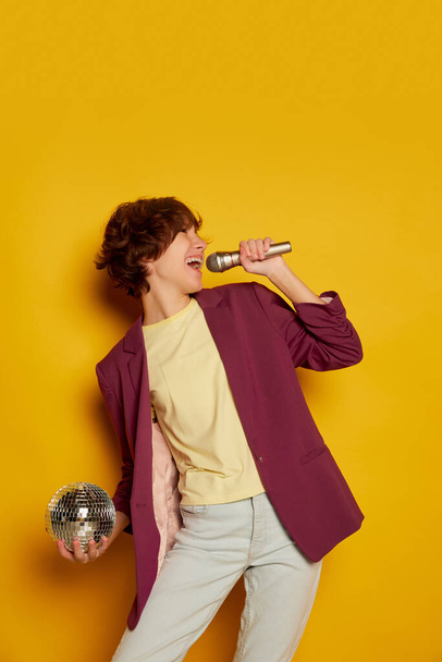Retrato de adolescente con pelo corto castaño rizado posando en chaqueta púrpura y cantando en micrófono aislado sobre fondo amarillo. Concepto de cultura juvenil, emociones, expresión facial, moda - Foto, Imagen