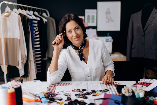 Porträt einer schönen kaukasischen Frau mit dunklen Haaren, die in einem modernen Atelier arbeitet. Reife Modedesignerin in stylischer Kleidung sitzt am Schreibtisch und zeichnet Papierskizzen. - Foto, Bild