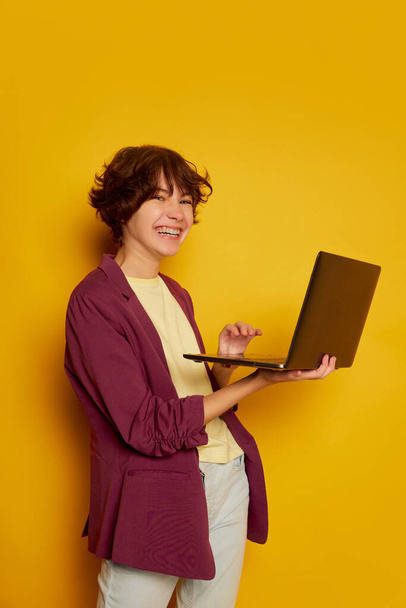 Portrait of cheerful teen girl, student with curly brown short hair posing with laptop isolated over yellow background. Concept of youth culture, emotions, facial expression, fashion - Fotoğraf, Görsel