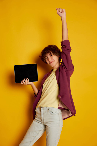 Portrait of cheerful teen girl with curly brown short hair posing with tablet isolated over yellow background. Online education. Concept of youth culture, emotions, facial expression, fashion - Foto, Imagen