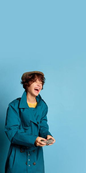 Portrait of cheerful teen girl with curly brown short hair posing in autumn coat and cap isolated over blue background. Concept of youth culture, emotions, facial expression, fashion - Foto, afbeelding
