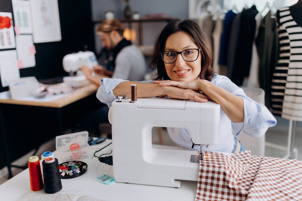 Aantrekkelijke mode-ontwerper in brillen leunend met hoofd op naaimachine en oprecht lachend. blank volwassen vrouw in bril werken op moderne tailoring atelier. - Foto, afbeelding