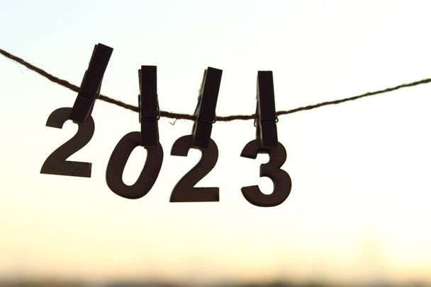 木製の番号は、オレンジ色の朝の空の日の出とぼやけた都市のボケ光の背景の上に服のペグピンで接続されたロープにハングアップします。明るいスタート日のコンセプトのための幸せな新年2023お祝い. - 写真・画像