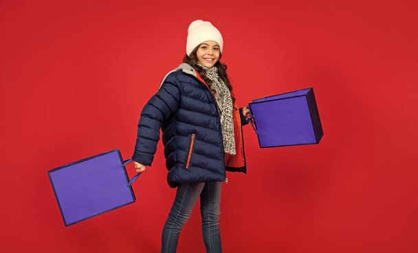 happy kid in puffer jacket and hat. teen girl after shopping on red background. child walking holding shopping bags with copy space. winter sales. - Фото, изображение