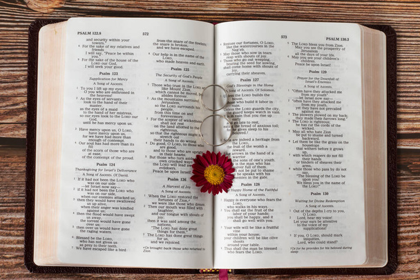 Otevřete biblickou knihu se dvěma svatebními prsteny a květinami. Pohled na horní stůl. Detailní záběr. Láska, věrnost, silný křesťanský vztah, a požehnání od Boha Ježíše Krista, biblický koncept. - Fotografie, Obrázek