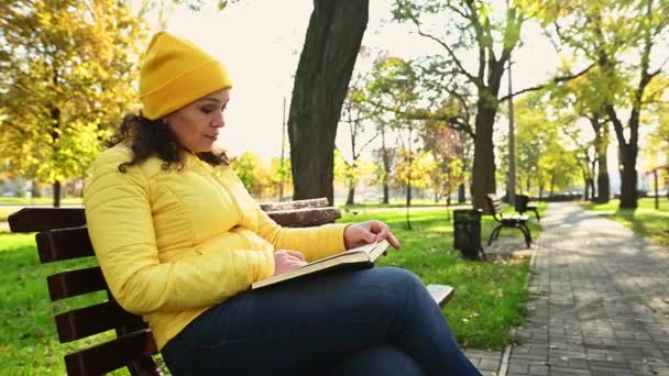 Charmante femme multi-ethnique dans une veste et un chapeau jaune vif, assise sur un banc en bois tout en se reposant dans le parc d'automne par une journée ensoleillée, livre de lecture. Désintoxication numérique, littérature. Loisirs. Été indien - Séquence, vidéo
