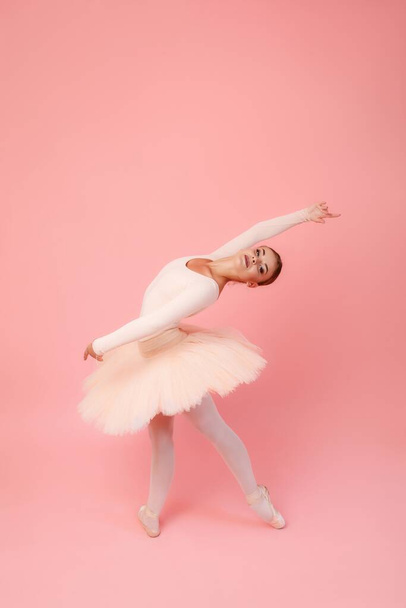 Hermosa bailarina flexible en tutú y zapatos puntiagudos doblando sobre fondo de estudio rosa. Encantadora joven que demuestra su gracia y profesionalidad - Foto, imagen