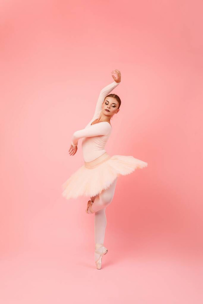 Aantrekkelijke jonge vrouw met flexibel lichaam die balletbewegingen uitvoert over roze studioachtergrond. Professionele ballerina met tutu en pointe schoenen. - Foto, afbeelding