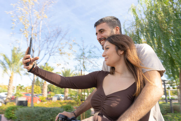 střední záběr pár milenců nahrávají sami jízdu na elektrickém skútru, zatímco procházejí městským parkem, usmívají se do kamery - Fotografie, Obrázek