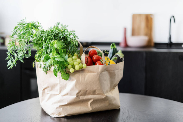bolsa de papel completa con verduras frescas, frutas y hierbas en la cocina casera con fondo borroso, dieta vegetariana, compra de comestibles y concepto de entrega - Foto, Imagen