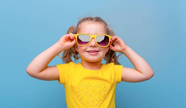 Little girl in yellow sunglasses on a blue background. - Photo, Image