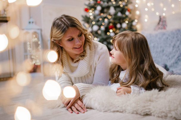 Familia Nochebuena feliz madre e hija en suéteres blancos se encuentran en la alfombra blanca en el fondo del árbol de Navidad y las luces. - Foto, Imagen