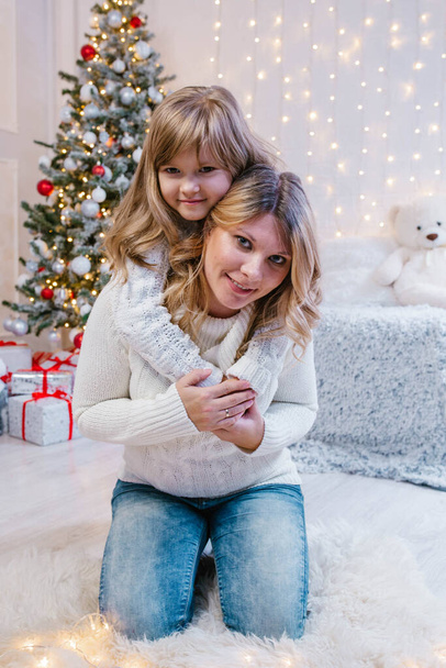 Családi karácsony este, boldog lánya ölelés anya hátulról ül térden, fehér pulóverben, a háttérben a karácsonyfa és fények. - Fotó, kép