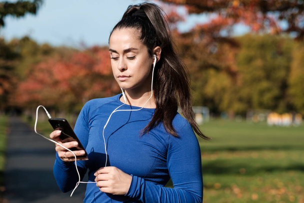 ヘッドフォンで音楽を聴くためにスマートフォンを使用してスポーツ女性。彼女は秋の晴れた日にロンドンの公園にいる。. - 写真・画像