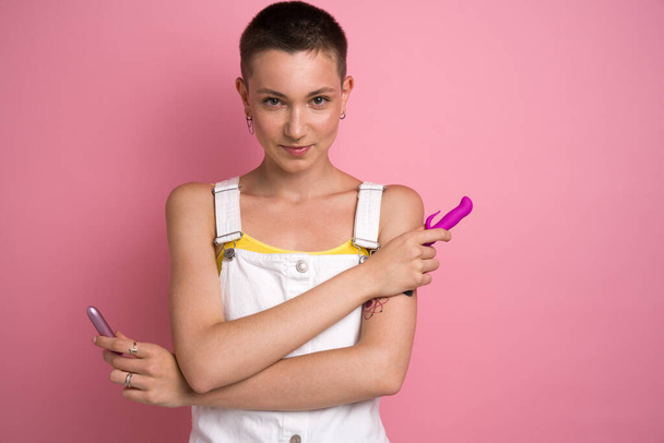 Sexy woman looking playfully at the camera while holding two dildo toys over the pink background. Excitement and fertility concept - Photo, Image