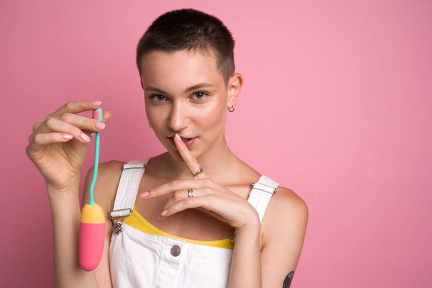 Cute shirt haired girl representing vibrating egg and holding finger near her lips over pink wall background. Equipment for satisfaction concept - Φωτογραφία, εικόνα