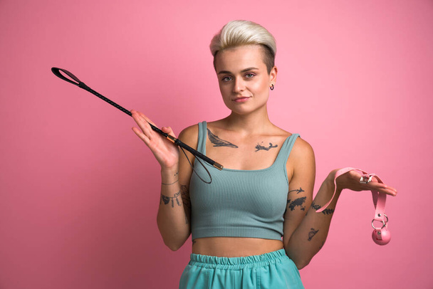 Positive dominant woman holding whip and gap, expresses passion and domination isolated on pink studio background. Sexual excitement concept - Valokuva, kuva