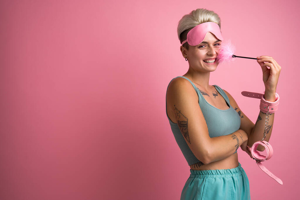 Cheerful tattooed woman wearing handcuffs laughing with pink feather stick while posing over isolated background. Stock photo - Foto, imagen